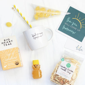 A gift box featuring items to uplift and comfort: a greeting card with the message "The Sun Will Come Up Tomorrow," cheerful Ray of Sunshine Lemondrops, comforting Chicken Noodle Soup (Serves 10), soothing Cup of Sunshine Tea Bags (10), a Mini Honey Bear, and Swizzle Sticks. Ideal for spreading warmth and positivity in a thoughtful gesture